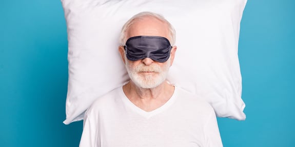 An elderly man rests on a white pillow with a black sleep mask covering his eyes.