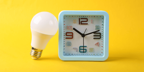 A white light bulb leaned up against a blue alarm clock on a yellow background