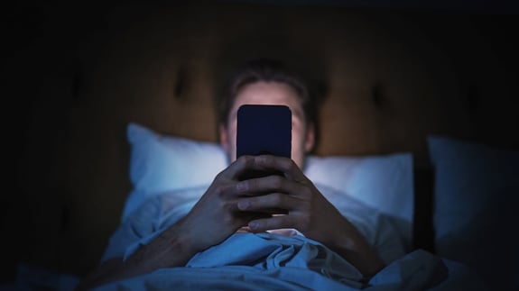 A man lies in bed with a phone in his hand, its glow illuminating his face