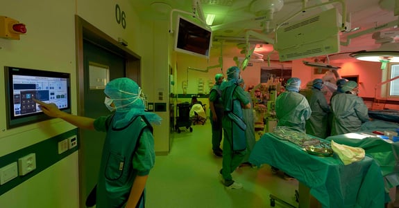 Nurse standing in the foreground presses a screen in an operating theatre with ergonomic lighting while the other staff is looking at a screen in the background