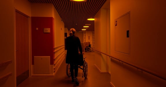 A healthcare worker takes an elderly citizen in a wheelchair down the corridor at a nursing home with circadian light from Chromaviso in the ceiling